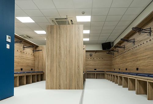 Modern empty locker room with wooden benches, coat hooks, and padded seating along walls.