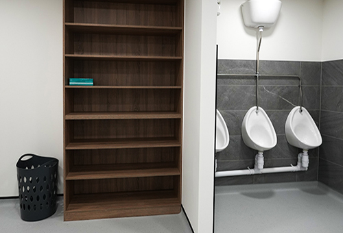 Clean restroom with three white urinals on gray tiled walls and an empty wooden shelf next to a laundry basket.