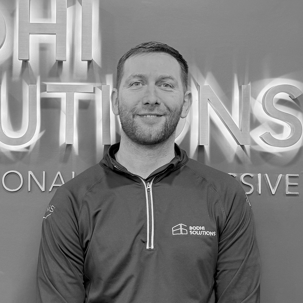 Smiling man in a Bodhi Solutions zip-up shirt standing in front of a lighted company sign.