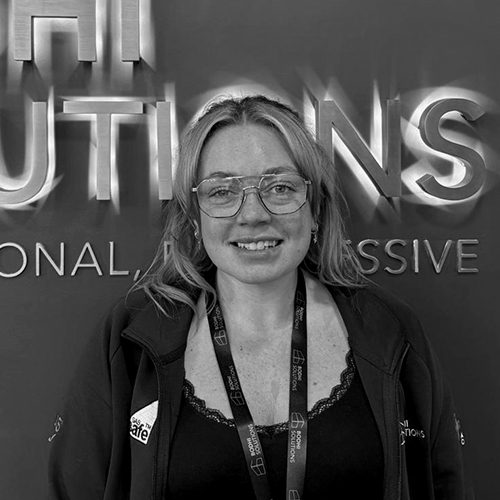 Smiling woman with glasses and lanyard stands in front of a sign with partial text Solutions.