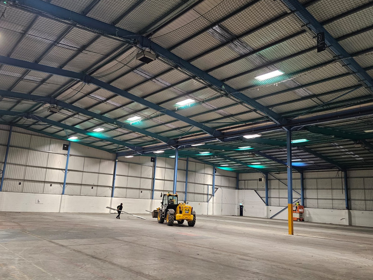 Large empty warehouse interior with a person and yellow construction vehicle on concrete floor.
