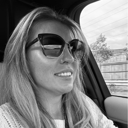 Woman with long hair wearing large sunglasses and smiling inside a car near a window.