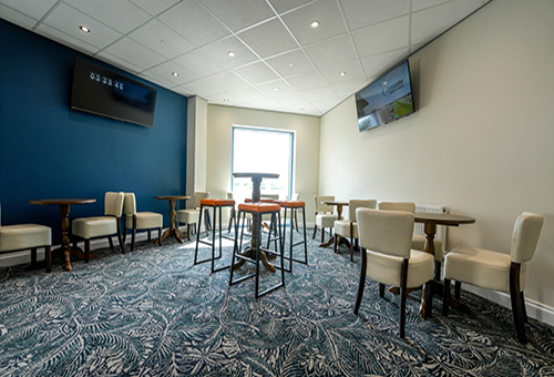 Modern meeting room with high tables, white chairs, patterned carpet, and two wall-mounted TVs.