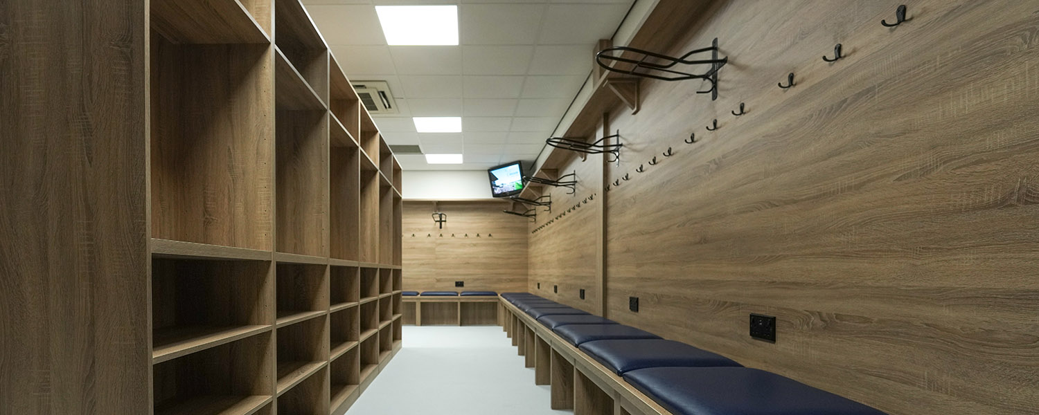 Modern locker room with wooden shelves, blue padded benches, coat hooks, and wall-mounted TV.