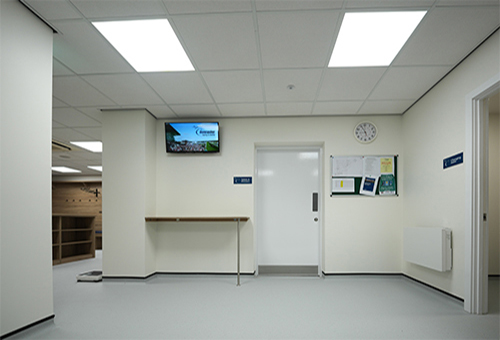 Clean, bright interior hallway with white walls, ceiling lights, bulletin board, and a digital screen on the wall.