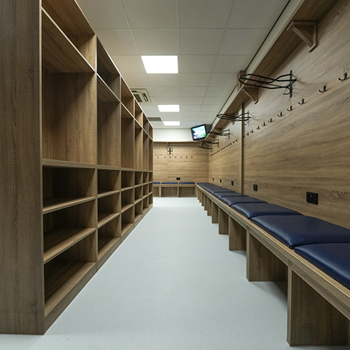 Empty locker room with wooden shelves, hooks, and blue cushioned bench seating along the walls.