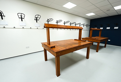 Empty white-walled room with wooden bench tables and black hooks mounted on walls.