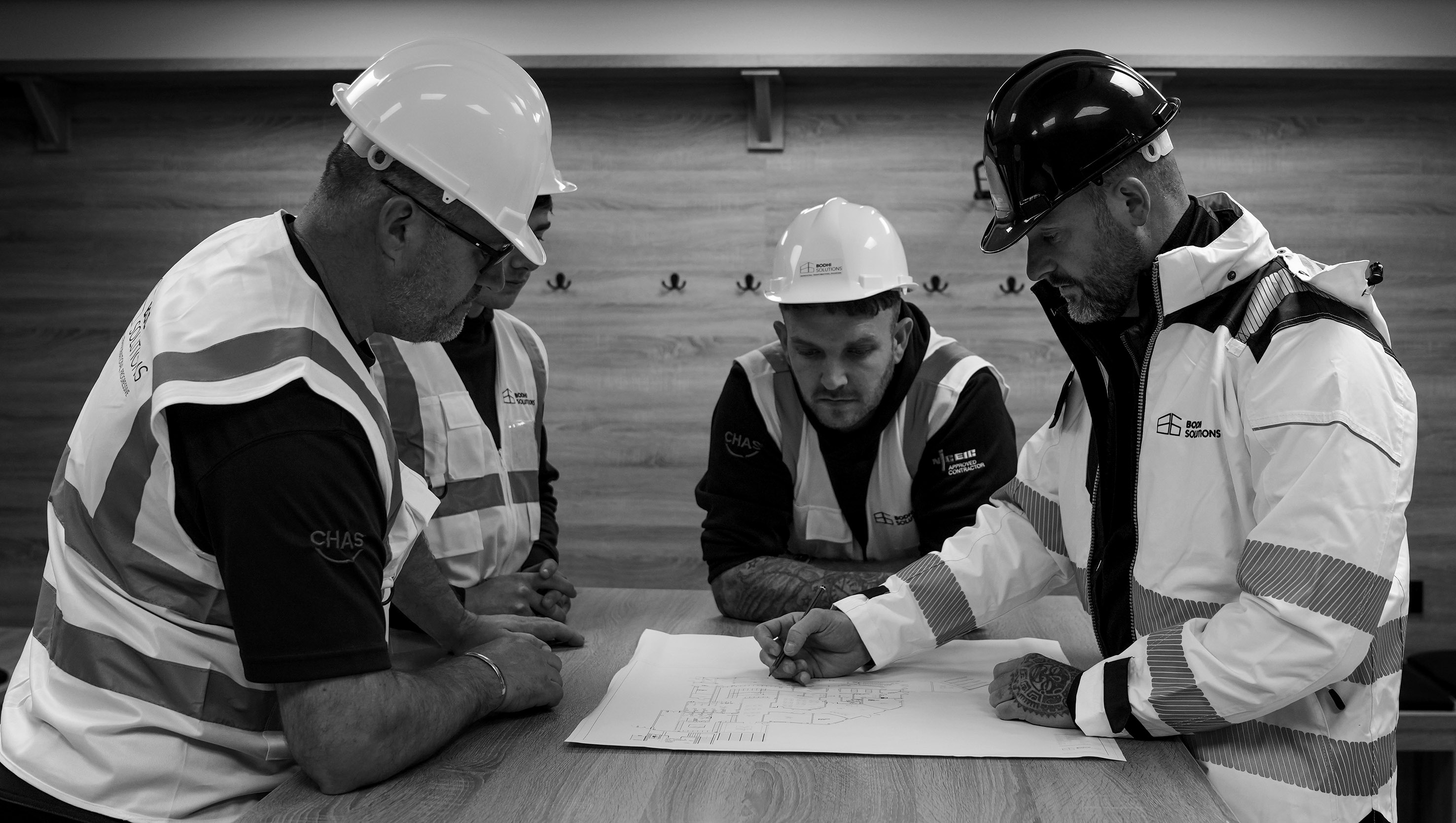 Four construction workers in helmets and vests review building plans on a wooden table.