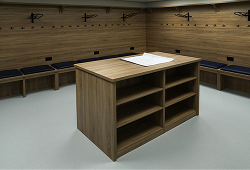 Empty locker room with wooden benches, black coat hooks, and a central wood storage unit with papers on top.