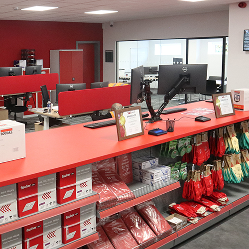 Modern office workspace with red desks, computer monitors, and shelves stocked with gloves and supplies.