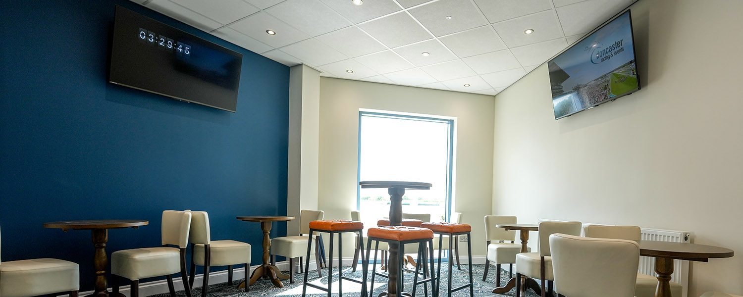 Modern seating area with cream chairs, wooden tables, blue wall, TV screens, and large window.
