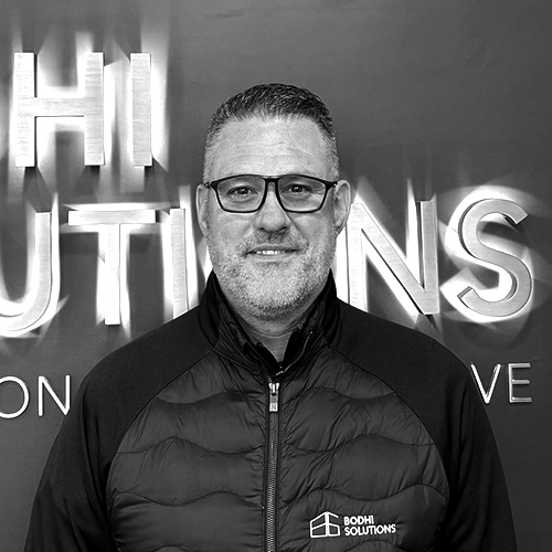 Man with glasses wearing a Bodhi Solutions jacket standing in front of a sign with large letters.