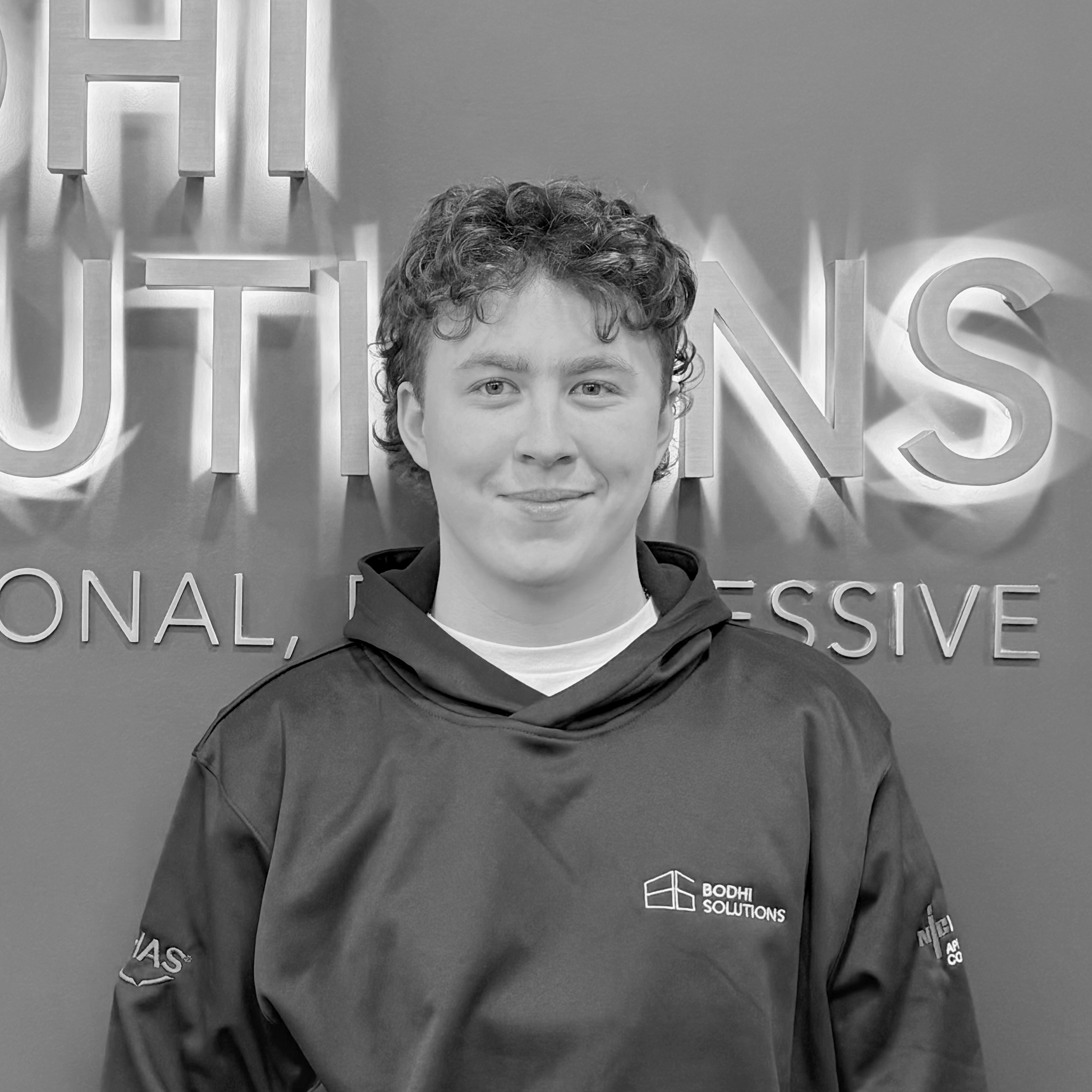 Young man with curly hair wearing a Bodhi Solutions hoodie, smiling in front of company sign.