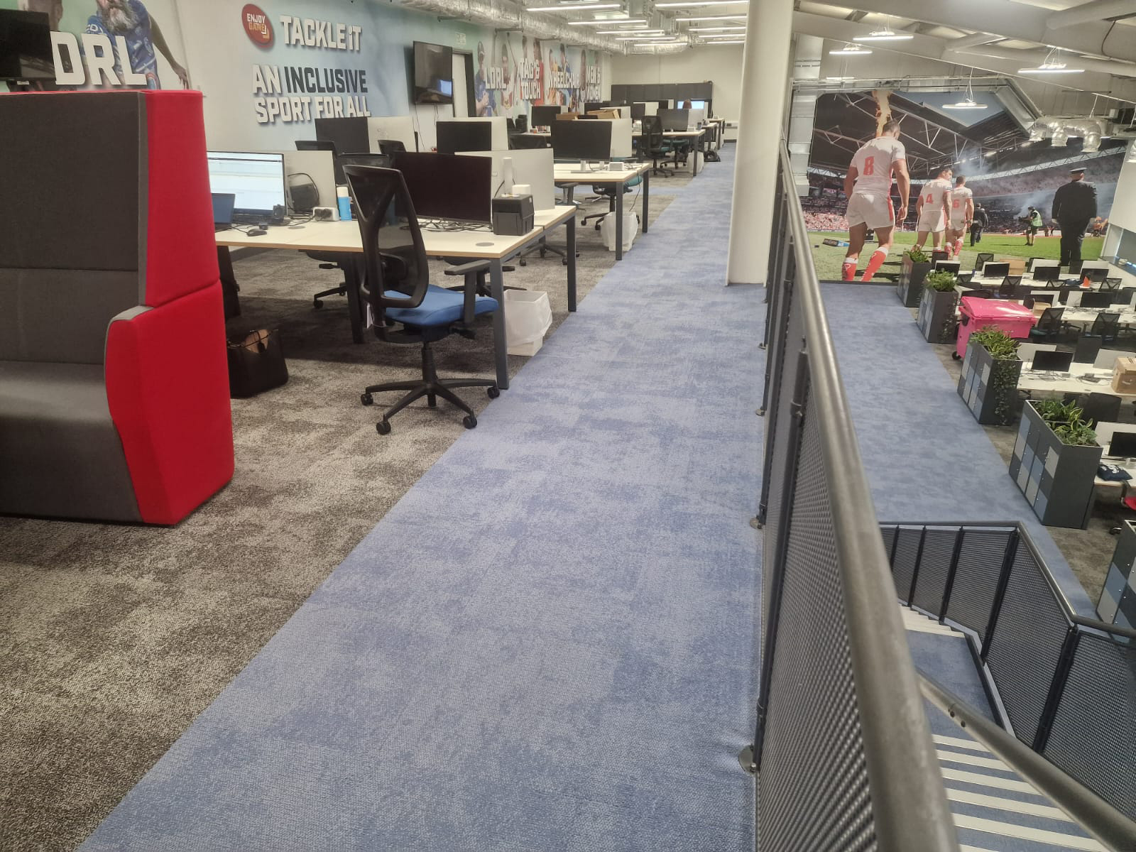 Open office with desks, chairs, blue carpet, and large sports-themed wall murals in a modern workspace.