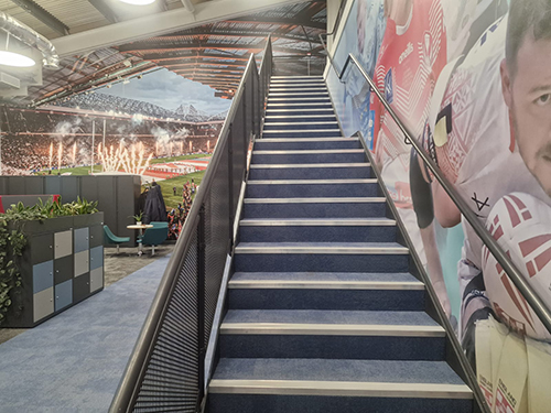 Indoor staircase with blue carpet and sports-themed murals and photos on walls inside a stadium lounge area.