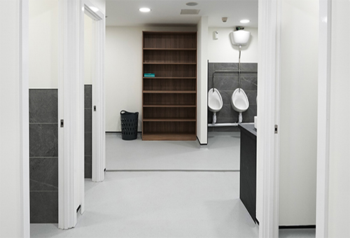 Minimalist commercial restroom with urinals, stalls, a wooden shelving unit, and a black laundry basket.