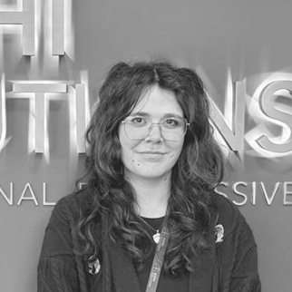 Woman with curly hair and glasses wearing a dark jacket stands in front of blurred large text.