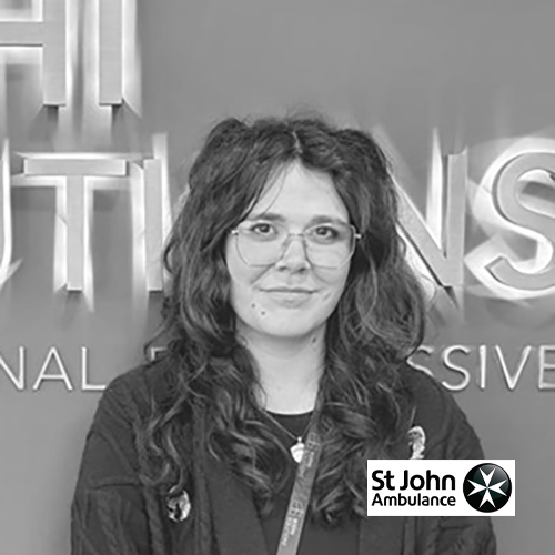 Young woman with long curly hair and glasses stands in front of a sign with St John Ambulance logo.