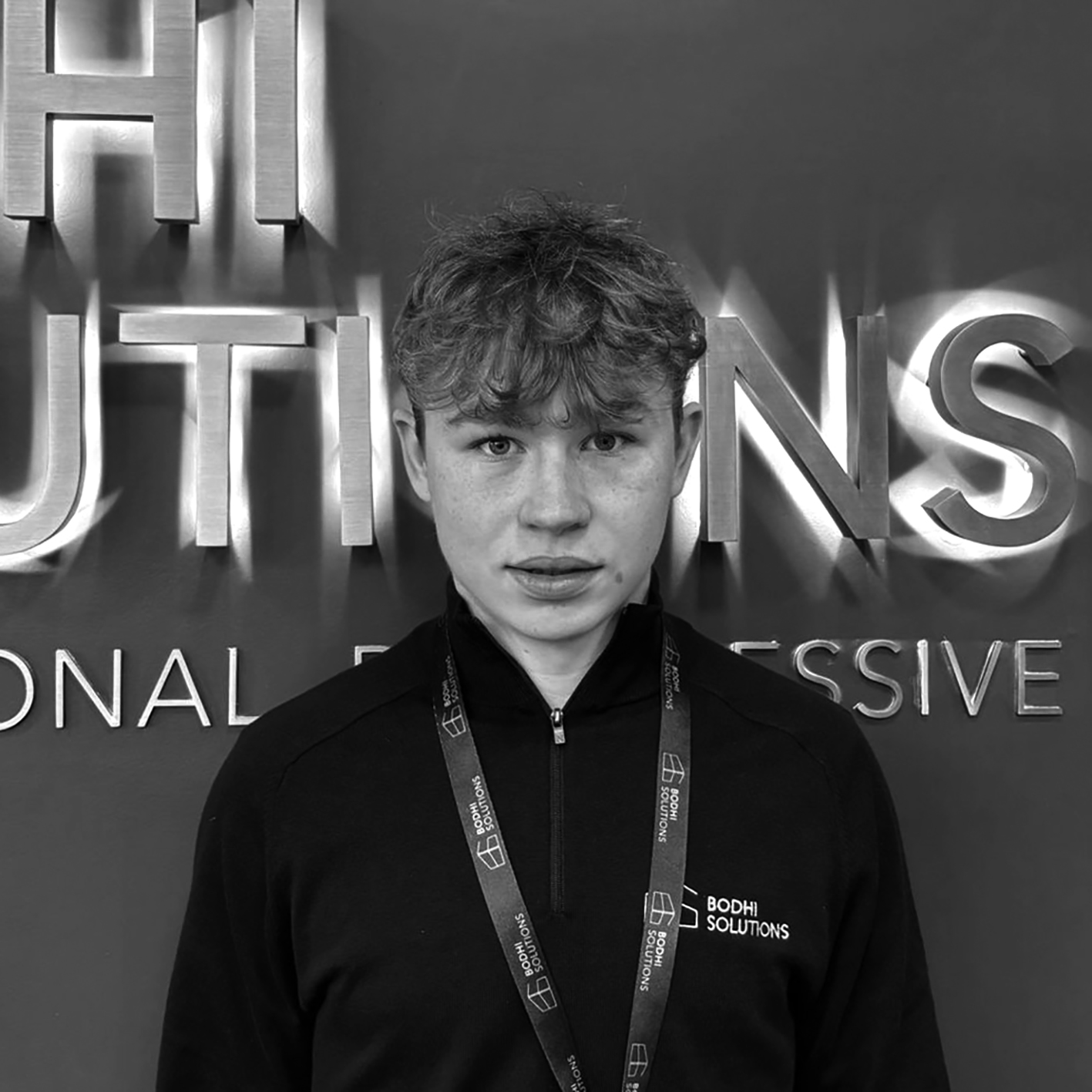 Teenage boy with curly hair wearing Bodhi Solutions lanyard and zip-up shirt in front of company sign.