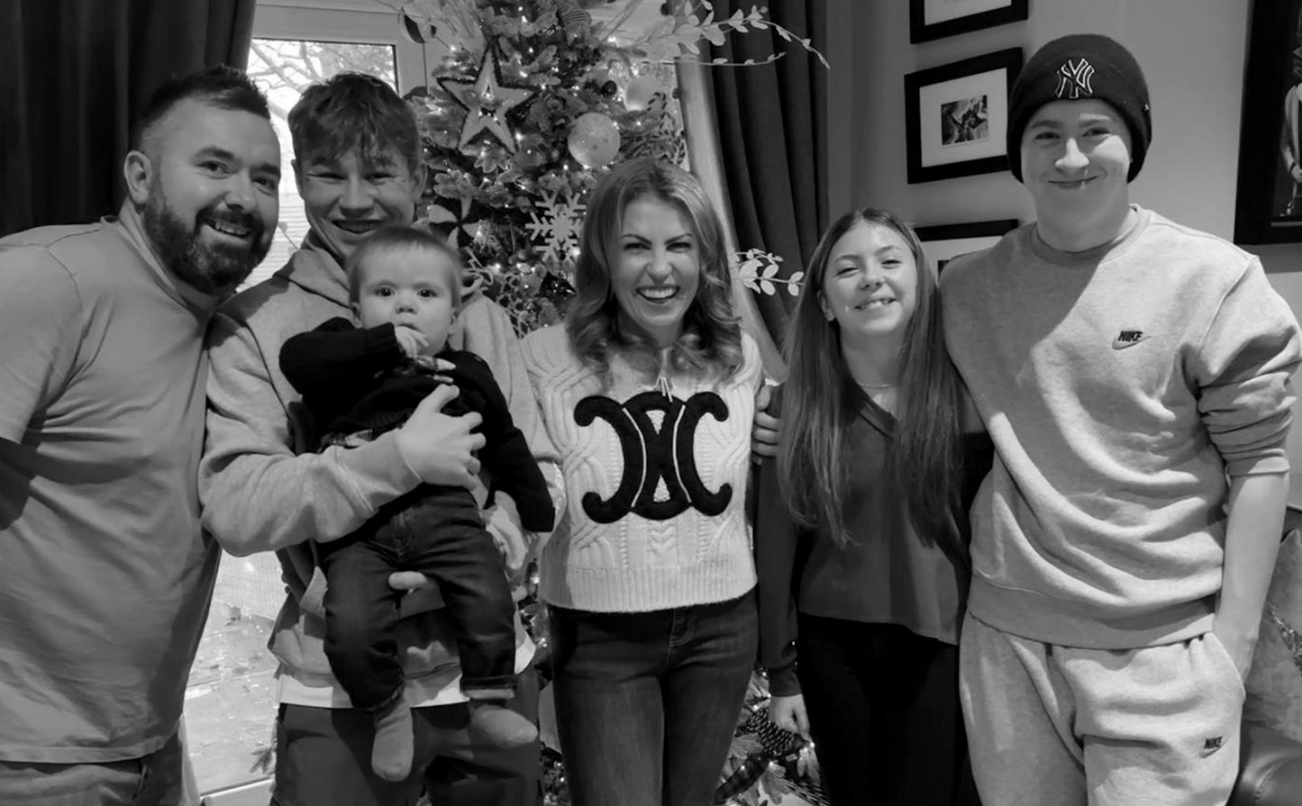 Smiling family of six, including a baby, posing in front of a decorated Christmas tree indoors.