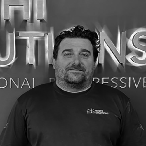 Man with dark hair and beard wearing a dark shirt with a company logo in front of a wall with large text.