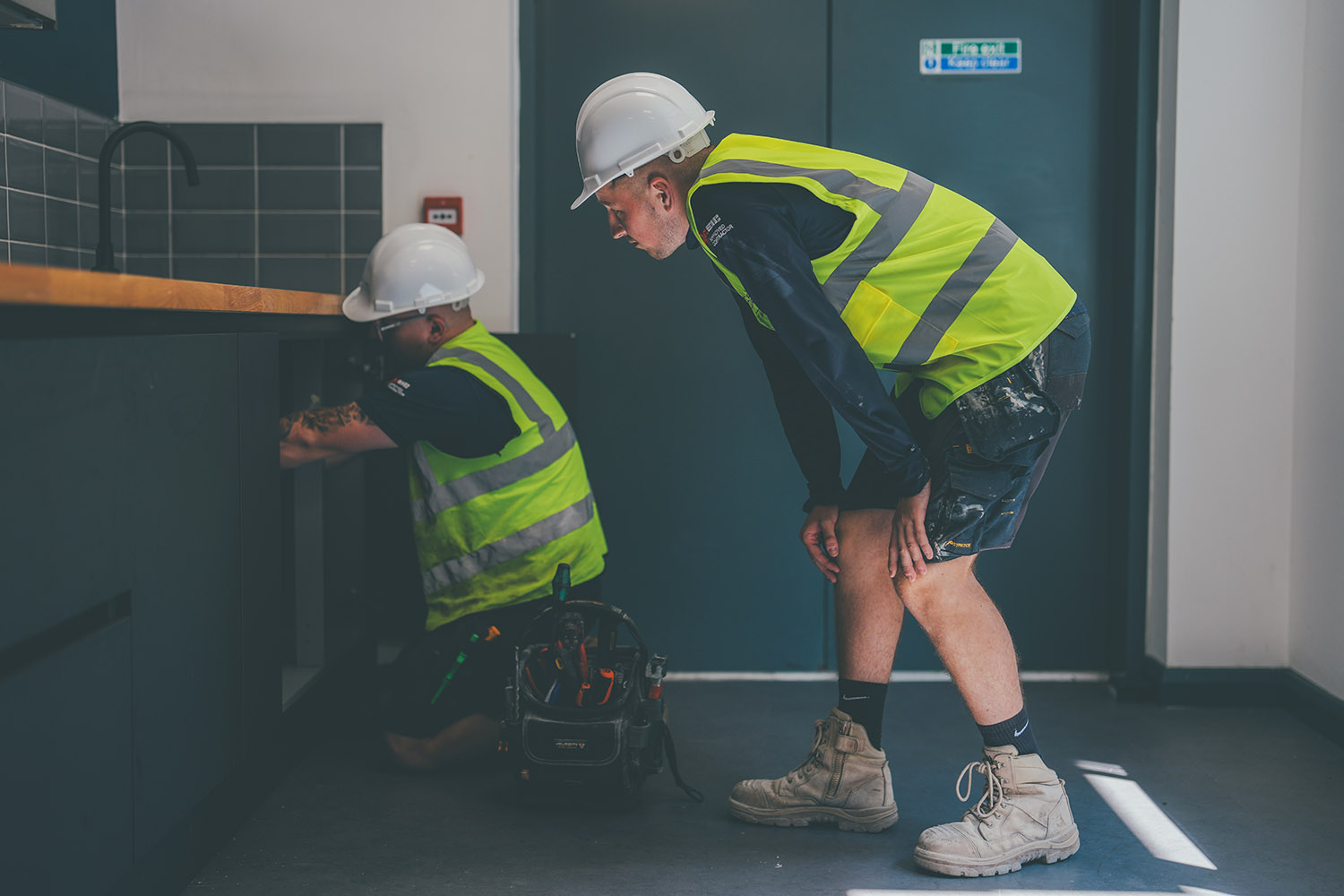 Two construction workers in yellow vests and white helmets working on cabinets indoors.
